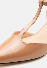 Μπεζ παπούτσι από λουστρίνι με μυτερή μύτη, T-λουράκι και λεπτομέρειες ομαλών ραφών σε λευκό φόντο.