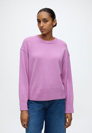 Vrouw met donker haar in een knot, die een losse roze trui met lange mouwen en blauwe jeans draagt, staand tegen een effen lichte achtergrond.