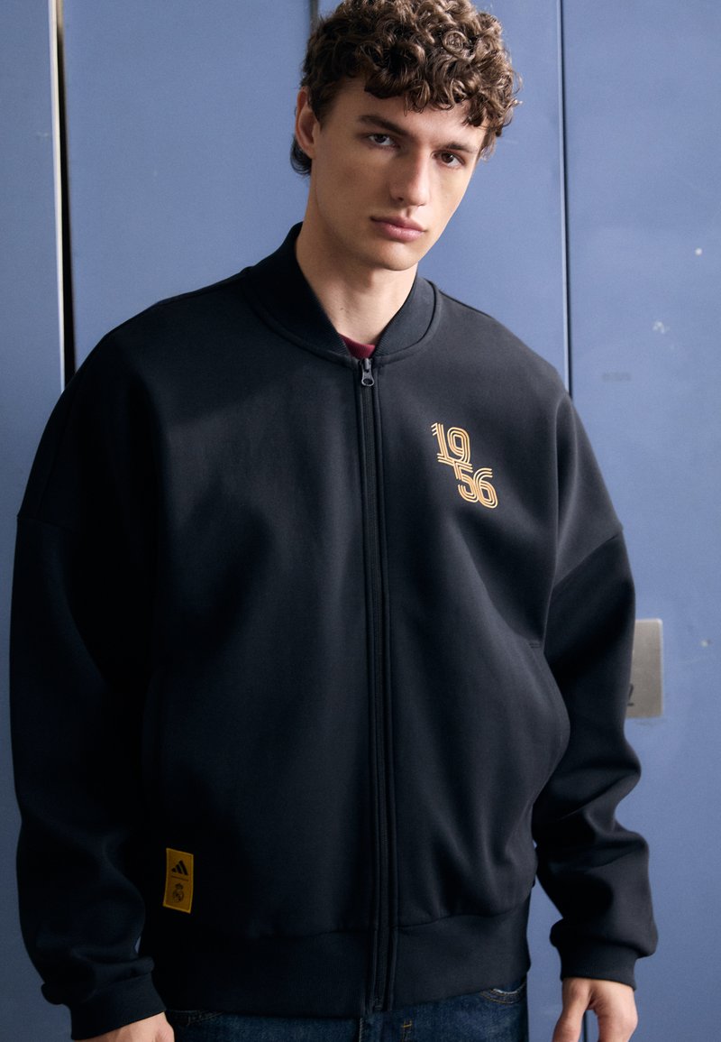 Black zip-up sweatshirt with a high collar, featuring gold “1956” embroidery and a yellow Adidas logo patch at the hem. Soft fabric, relaxed fit.