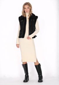 Zwarte leren vest met nepbontvoering en ritssluiting, gecombineerd met een crèmekleurige ribgebreide midi-jurk en zwarte hoge laarzen tot de knie.