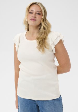 Blondynka o długich włosach, ubrana w biały top z falbanami na rękawach i niebieskie jeansy, stoi na tle jasnoszarego, jednolitego tła.