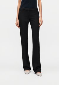 Γυναίκα που φοράει μαύρο, ραμμένο παντελόνι με ίσια γραμμή και μπεζ ψηλοτάκουνα παπούτσια με μυτερή μύτη, στέκεται μπροστά σε απλό λευκό φόντο.