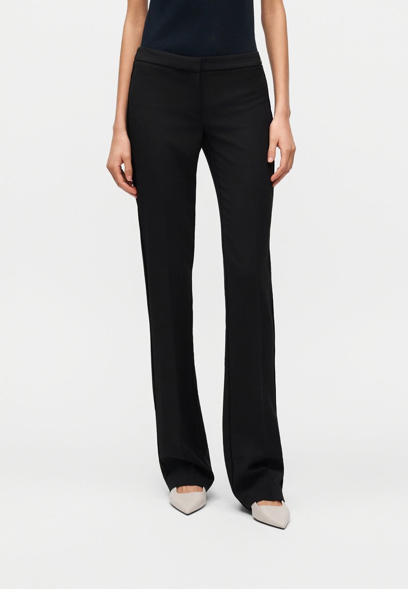 Γυναίκα που φοράει μαύρο, ραμμένο παντελόνι με ίσια γραμμή και μπεζ ψηλοτάκουνα παπούτσια με μυτερή μύτη, στέκεται μπροστά σε απλό λευκό φόντο.