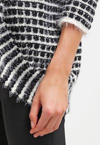Pull en rayures noir et blanc avec une texture pelucheuse. Présente des manches larges et amples et un ourlet irrégulier. Une main tient le bord.