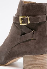 Bottine en daim marron avec une boucle dorée et un détail de bride croisée, dotée d'un talon en bois empilé et d'une texture lisse.