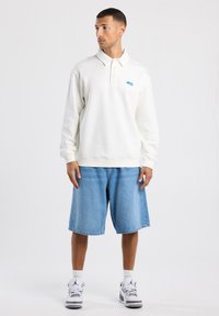 Felpa bianca a polo con colletto e pattina dei bottoni, caratterizzata dal logo "NYC" in blu. Abbinata a pantaloni corti in denim azzurro e scarpe da ginnastica.