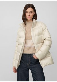 Žena v krémovej prešívanej bunde, ktorá má na sebe béžový sveter a tmavé džínsy, pózuje s jednou rukou v vrecku na jednotnom pozadí.