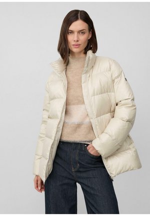 Kobieta w kremowej puchowej kurtce na beżowym swetrze i ciemnych dżinsach, pozująca z jedną ręką w kieszeni na jednolitym tle.