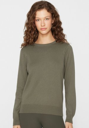 Jeune femme aux cheveux bouclés portant un pull en maille à manches longues de couleur vert olive, debout devant un fond clair uni.