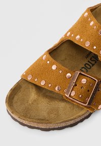 Καφέ σουέντ σανδάλι με δύο ρυθμιζόμενες λωρίδες που διαθέτουν μεταλλικά καρφιά και αγκράφα σε χάλκινο χρώμα. Ανατομική σόλα με υφή.