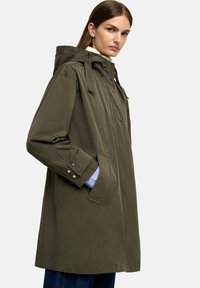 Oliwkowa wodoodporna parka z kapturem, z zamkiem błyskawicznym z przodu, z zapięciem na guziki i kieszeniami bocznymi. Luźny krój z regulowanymi trokami.