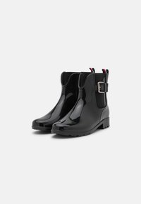 Bottes de pluie noires brillantes à cheville avec des panneaux élastiques latéraux, une boucle décorative et des poignées rouges-blanches-bleues sur un fond blanc.
