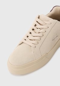 Beżowe zamszowe sneakersy z płaskimi sznurówkami, teksturowaną cholewką i niskoprofilową gumową podeszwą. Zawierają subtelne logo po bokach.