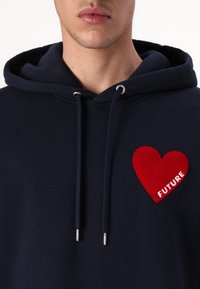 Felpa con cappuccio blu navy che presenta un patch a forma di cuore rosso con la parola "FUTURE" in testo bianco. Texture morbida con dettagli in occhio di bue argentati.
