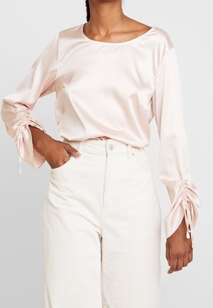 Personne portant un haut en satin rose clair à manches longues avec poignets froncés et un pantalon blanc en velours côtelé taille haute, main dans la poche.
