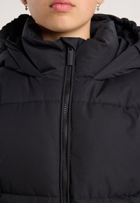 Persona que lleva una chaqueta acolchada negra con cremallera frontal cerrada hasta el cuello y pequeños pendientes de aro dorados visibles.