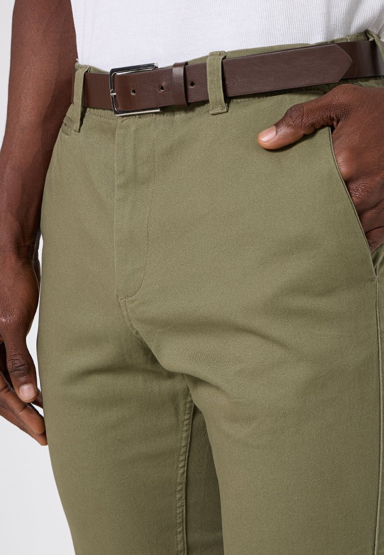 Olivgrön bomullsbyxor med rak skärning, som har sidofickor och ett brunt läderbälte med en silverspänne. Slät textur.