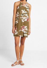 Olivgrünes Top-up-Dress mit floralem Muster in Rot und Weiß, mit einem Schlitz-Ausschnitt und fließendem Design. Kombiniert mit silbernen Sandalen.