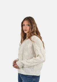 Maglione bianco lavorato a maglia con un design a trecce testurizzato; presenta maniche lunghe e un scollo rotondo, abbinato a jeans blu.