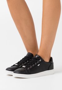 Une paire de baskets noires texturées avec des semelles blanches portées pieds nus sur un fond blanc.