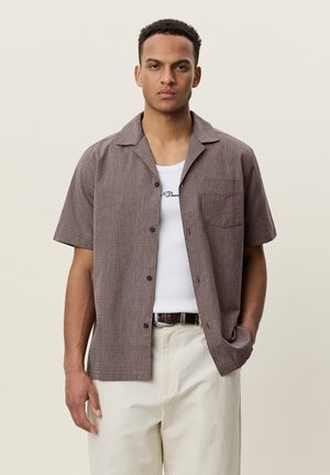 Homme portant une chemise marron à manches courtes boutonnée par-dessus un débardeur blanc et un pantalon beige clair, debout devant un fond uni.