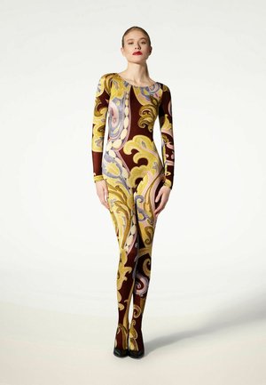 Donna in piedi che indossa una tuta aderente a maniche lunghe con motivo astratto a spirale giallo, marrone e viola e scarpe nere appuntite.