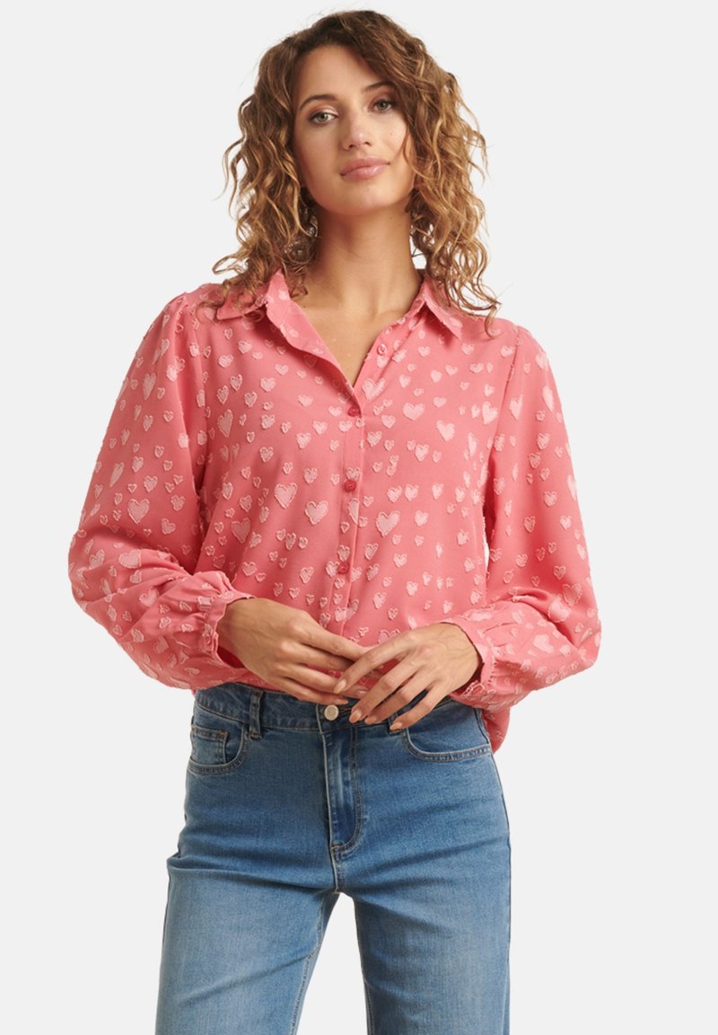 Chemise rose à boutons avec des motifs de cœurs subtils, manches longues avec des poignets élastiques, accompagnée de jeans bleus taille haute.