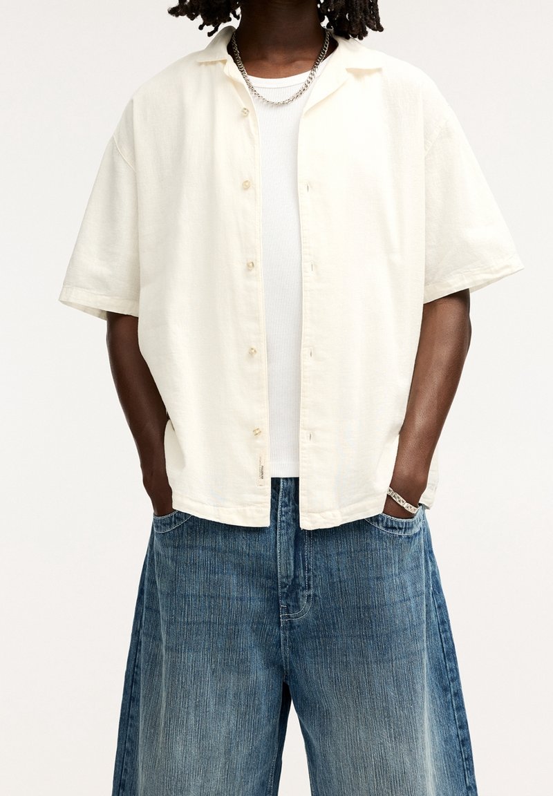 Lichtbeige kortermelig overhemd met knopen, gemaakt van een gestructureerde stof, gecombineerd met losse, vervaagde blauwdenim shorts met een wijde pijp.