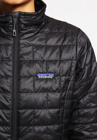 Schwarze gesteppte Jacke mit hohem Kragen, ausgestattet mit einem vollständigen Reißverschluss und einem kleinen Patagonia-Logo auf der Brust. Glatte, glänzende Stofftextur.