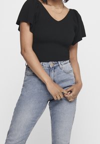 Top negro de canalé con mangas cortas y volantes, combinado con vaqueros de cintura alta de color azul claro. Capa notable de collares delicados.