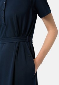 Granatowa sukienka polo z dopasowaną talią z wstążką, krótkimi rękawami, zapięciem na guziki i kieszeniami po bokach. Teksturowany materiał z akcentem w postaci logo.