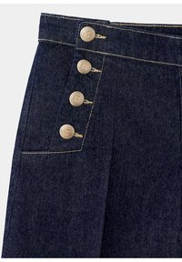 Pantalon en jean bleu foncé avec quatre boutons décoratifs en laiton sur le côté gauche à la taille et la couture adjacente.