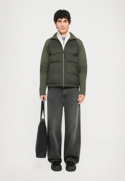 Young man in green puffer jacket, gray zip-up underneath, wide black jeans, black shoes, holding black tote bag, standing against white background.