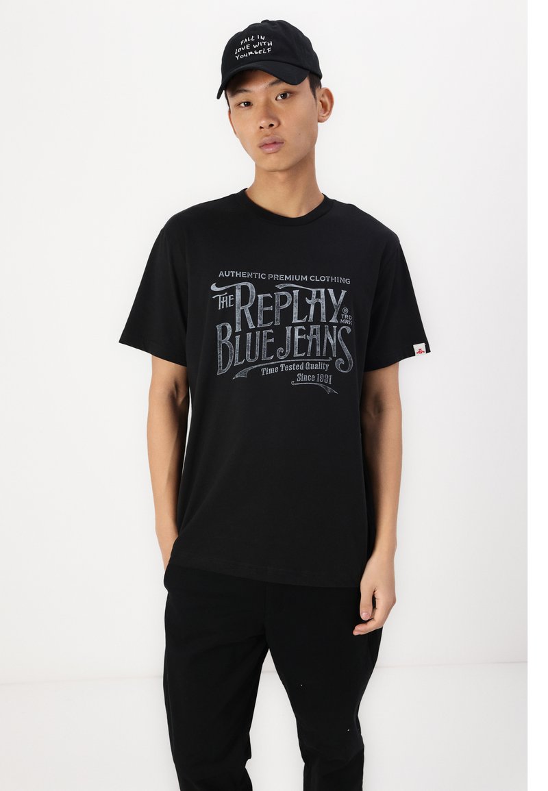 Schwarzes Baumwoll-T-Shirt mit einem Grafikdruck in Hellblau, auf dem der Text "THE REPLAY BLUE JEANS" auf der Vorderseite zu sehen ist. Getragen mit schwarzen Hosen.