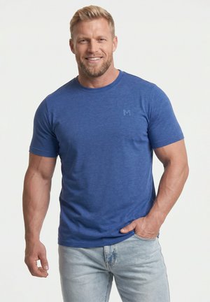 Uśmiechnięty muskularny mężczyzna z krótkimi blond włosami, ubrany w niebieski t-shirt i jasne dżinsy, z jedną ręką w kieszeni, na jednolitym jasnym tle.