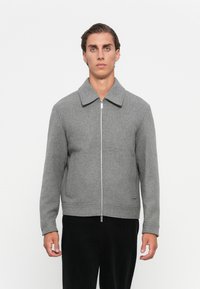 Ung mann står, iført en grå zip-up jakke med krage og svarte bukser, mot en ensfarget hvit bakgrunn.