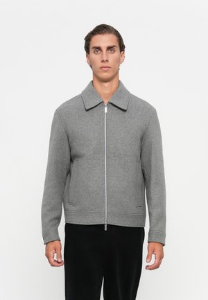 Young man standing, wearing a gray zip-up jacket with a collar and black pants, against a plain white background.
