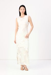 Robe longue sans manches en tissu transparent, ornée de broderies florales et dotée d'une fente sur le côté. Sandales légères de couleur marron clair avec des lanières.