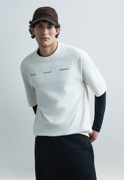 White cotton short-sleeve t-shirt with black text; layered over a black long-sleeve shirt. Brown baseball cap. Simple geometric design.
