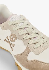 Sneaker featuring a beige suede overlay and cream fabric, with a skull and crossbones design, textured laces, and a rubber sole.