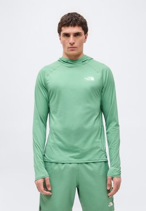 Muž v svetlozelenom dlhom rukáve s kapucňou a zodpovedajúcich šortkách s logami The North Face, stojaci pred jednoduchým bielym pozadím.