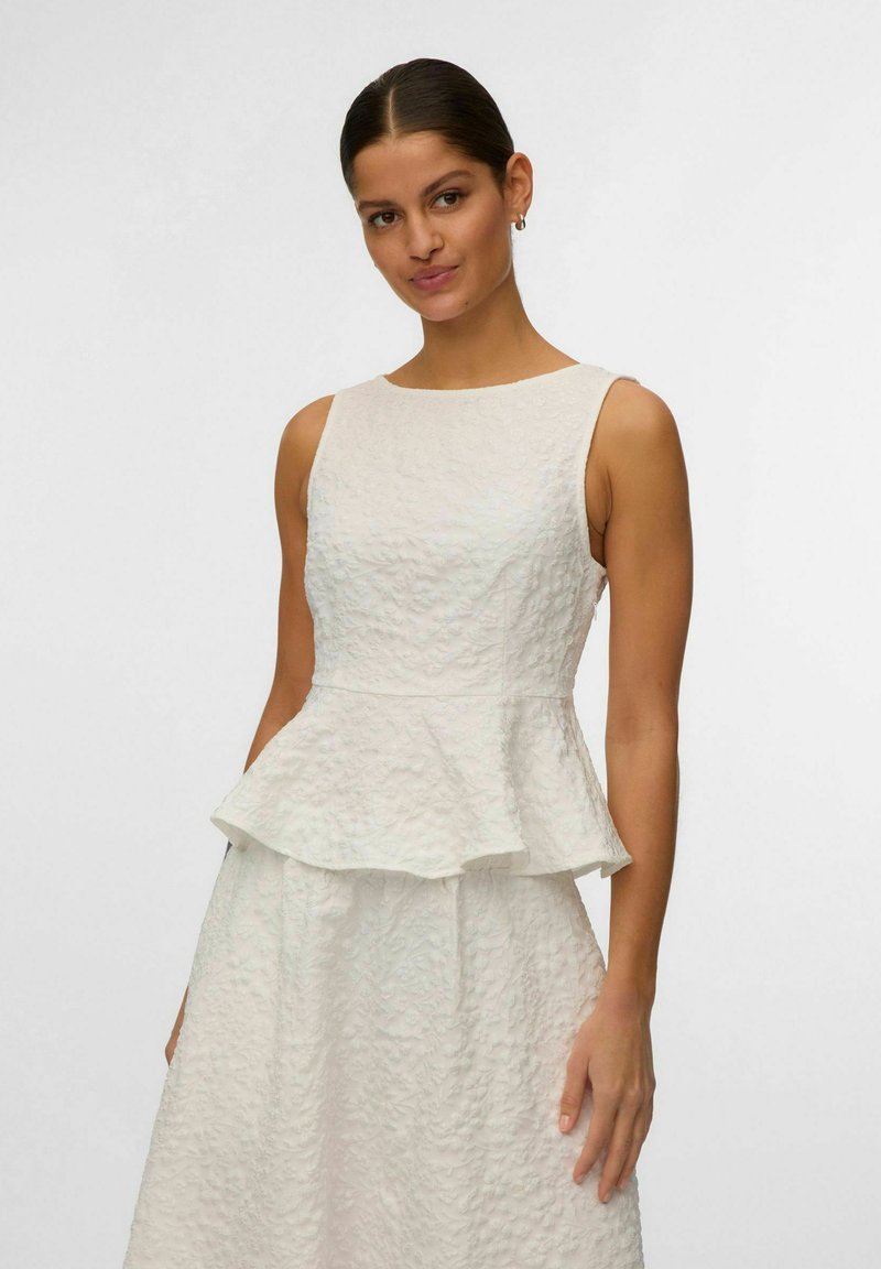 Femme aux cheveux foncés portant un haut péplum blanc texturé sans manches et une jupe assortie, sur un fond clair uni.