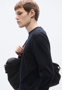 Črna ramenska torbica z zategovalnim vrvico, nošena na ramenu; model v temni majici z dolgimi rokavi z okroglim izrezom in subtilnim logotipom.