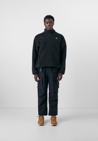 Schwarzer Fleece-Pullover mit hohem Kragen, Reißverschluss und dezentem Logo. Kombiniert mit schwarzen Cargo-Hosen und beigen Sneakers.