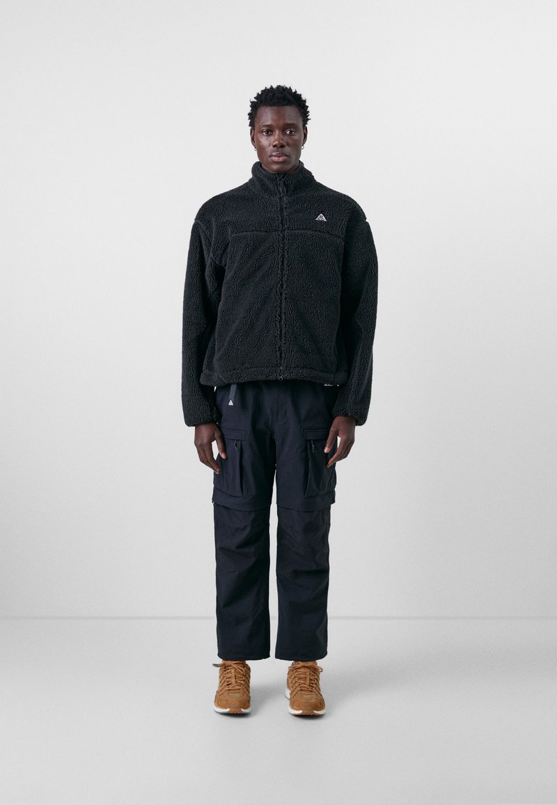 Schwarzer Fleece-Pullover mit hohem Kragen, Reißverschluss und dezentem Logo. Kombiniert mit schwarzen Cargo-Hosen und beigen Sneakers.