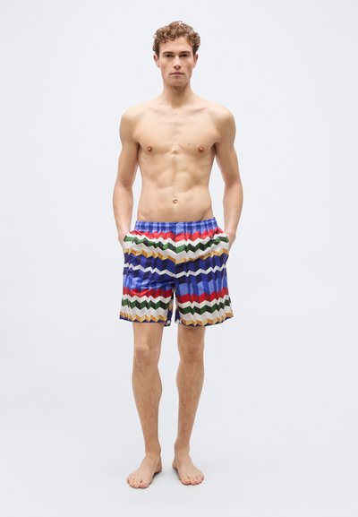 Νεαρός άνδρας όρθιος, ξυπόλυτος με τα χέρια στις τσέπες, φορώντας πολύχρωμα μαγιό με γεωμετρικά σχέδια πάνω σε απλό φωτεινό φόντο.