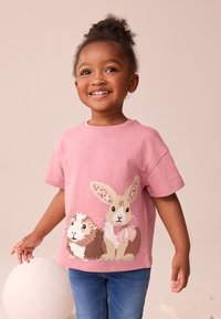 T-shirt rose clair à manches courtes avec un motif de lapin et de cochon d'Inde orné d'accents floraux. Porté avec un jean en denim bleu.