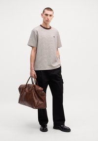 Pasiasta koszula z krótkim rękawem w kolorze beżowym i brązowym, czarne spodnie oraz duża brązowa torba podróżna ze skóry z dwoma uchwytami. Czarne buty.