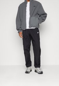 Giacca in fleece grigia con zip frontale, tasche grigie, t-shirt bianca sotto. Pantaloni cargo neri e sneakers grige con suola testurizzata.