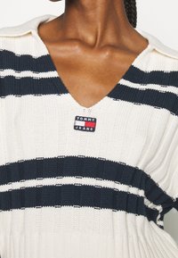 Clavicule et cou d'une personne portant un pull en maille crème à rayures horizontales bleu marine avec un patch logo Tommy Jeans au centre de la poitrine.